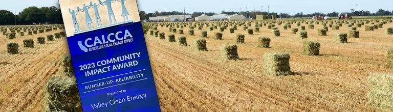 A blue community impact award sign stands in front of a field with neatly stacked hay bales under a clear sky.