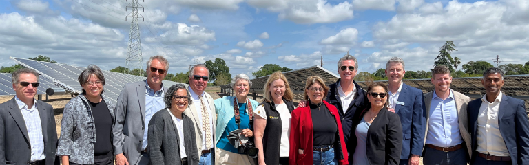 A group of people stands outdoors in front of solar panels, smiling at the camera under a partly cloudy sky.