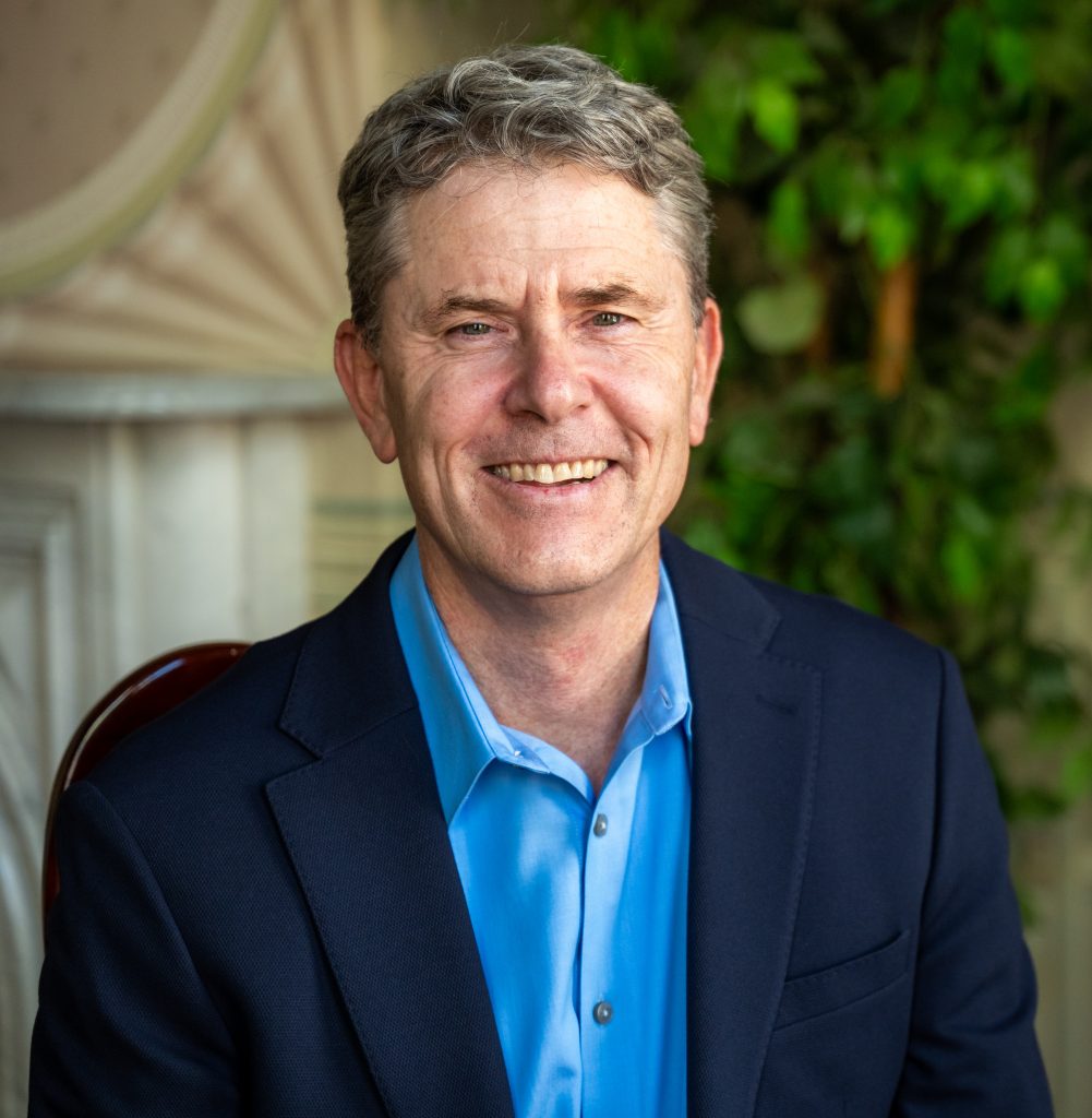 Man in a blue dress shirt and dark blazer smiling while seated indoors, with a leafy green background.