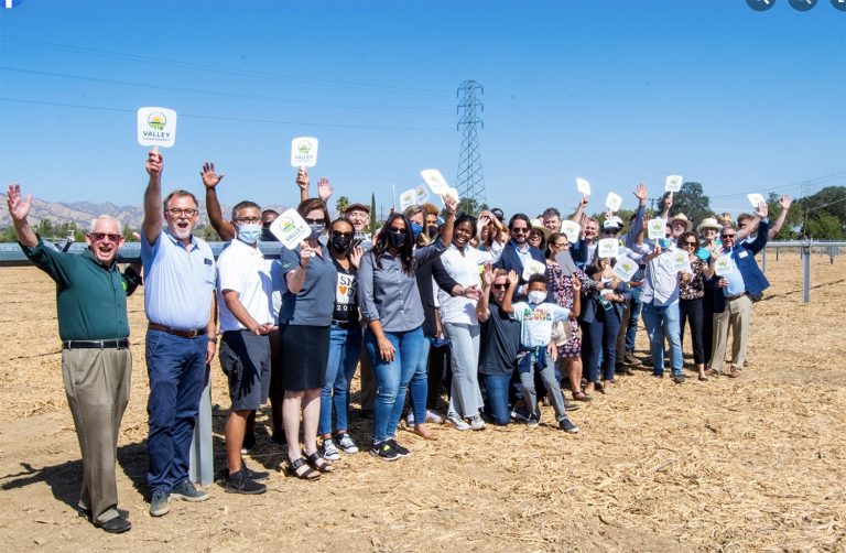 A group of adults and children stand outdoors on dirt, smiling and holding up signs that read Valley Clean Energy under a clear sky.