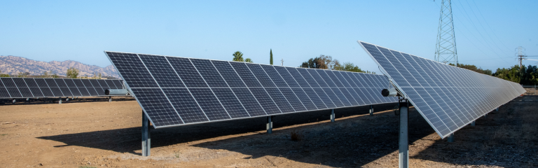 Rows of solar panels installed outdoors on a clear day, with mountains and power lines visible in the background.