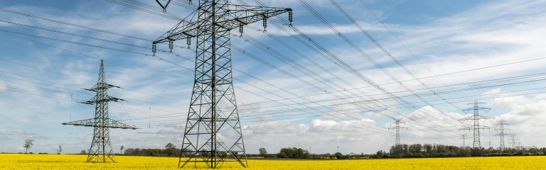 High-voltage power lines and towers stretch across a yellow field under a partly cloudy blue sky.