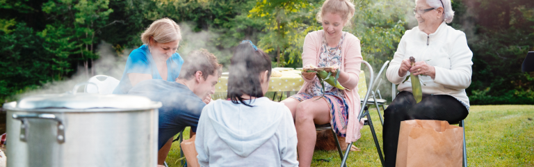 A group of people sit outdoors by a table and steam pot, preparing vegetables together on a grassy lawn.
