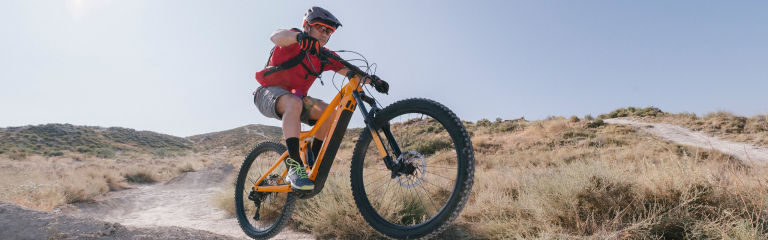 A person wearing a helmet rides a mountain bike on a dirt trail through dry, grassy hills under a clear sky.