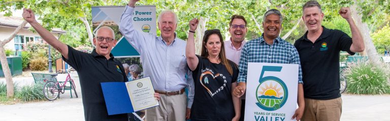 A group of six people celebrate Valley Clean Energys 5 years of service, holding a plaque and a sign, all smiling outdoors.