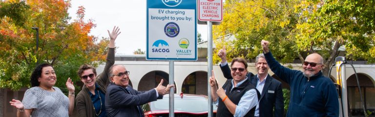 Seis personas posan junto a unas señales de recarga para vehículos eléctricos recién instaladas, sonriendo y posando para una foto de grupo al aire libre.