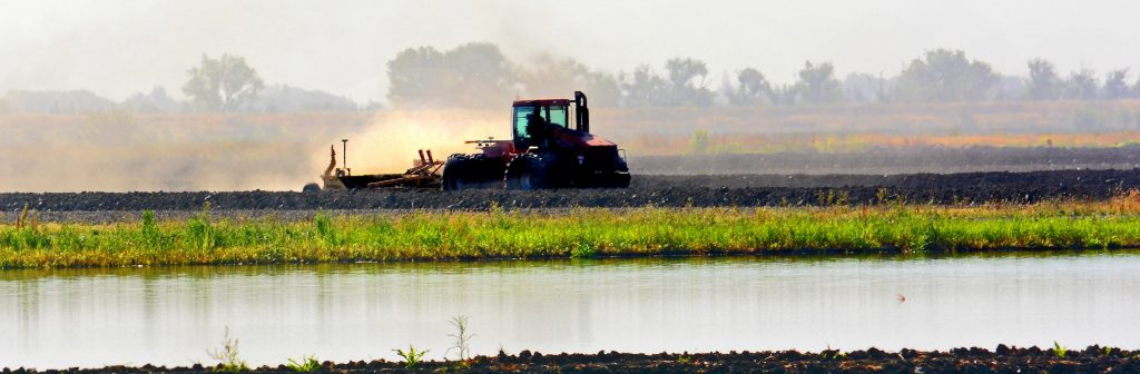 A red tractor plows a field near a body of water, with trees and fields visible in the background under a hazy sky.