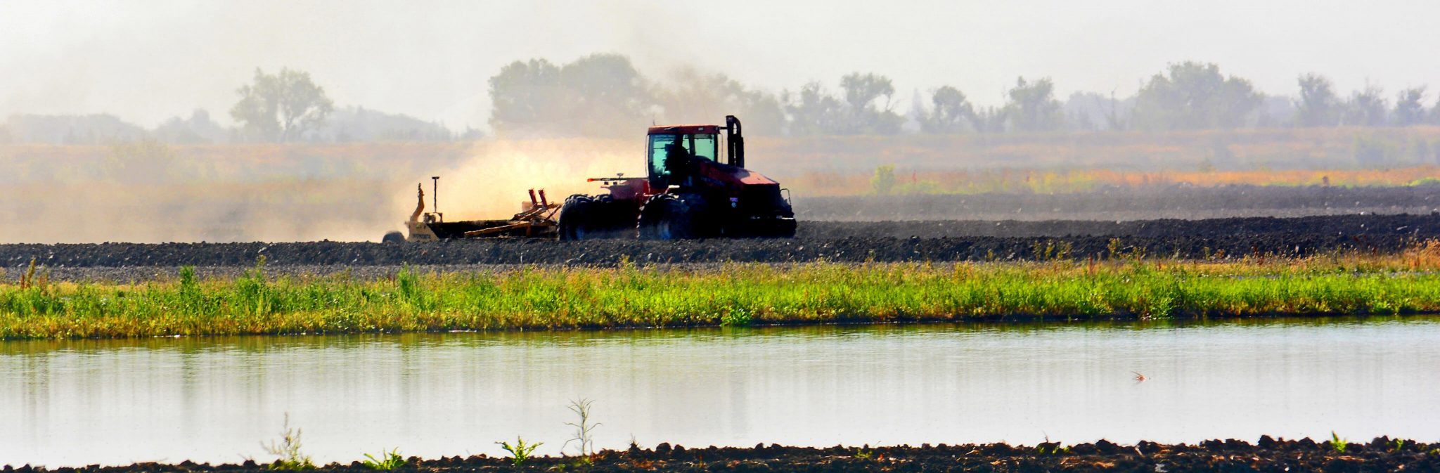 A red tractor plows a field near a body of water, with trees and fields visible in the background under a hazy sky.