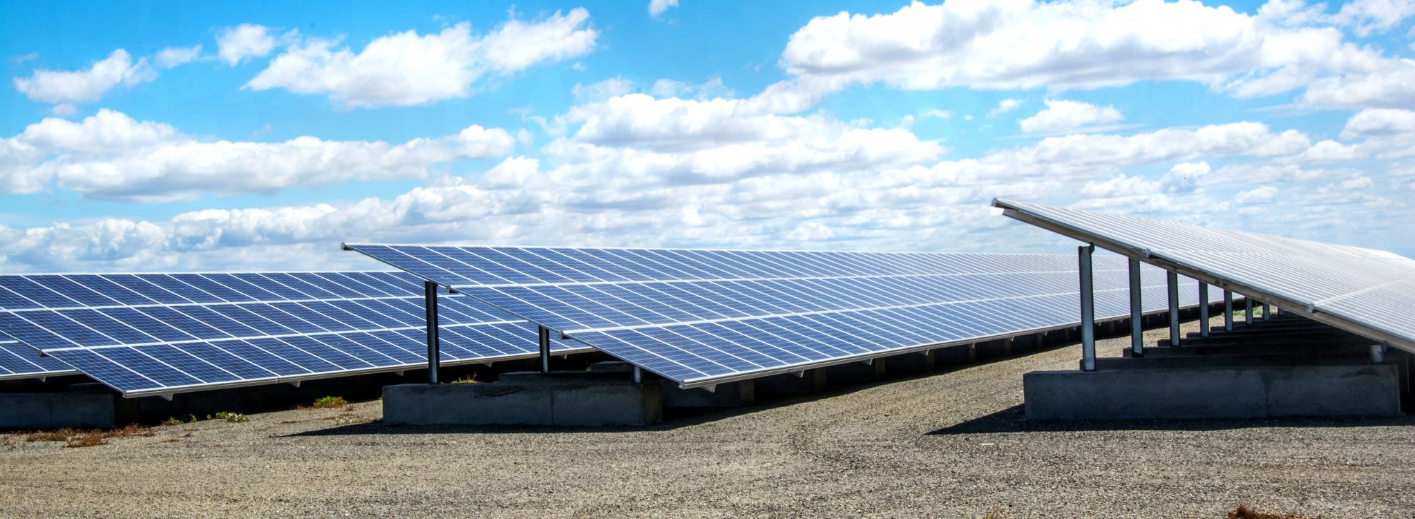 Rows of solar panels installed outdoors on a gravel surface under a partly cloudy sky.