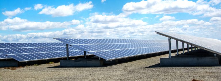 Rows of solar panels installed outdoors on a gravel surface under a partly cloudy sky.