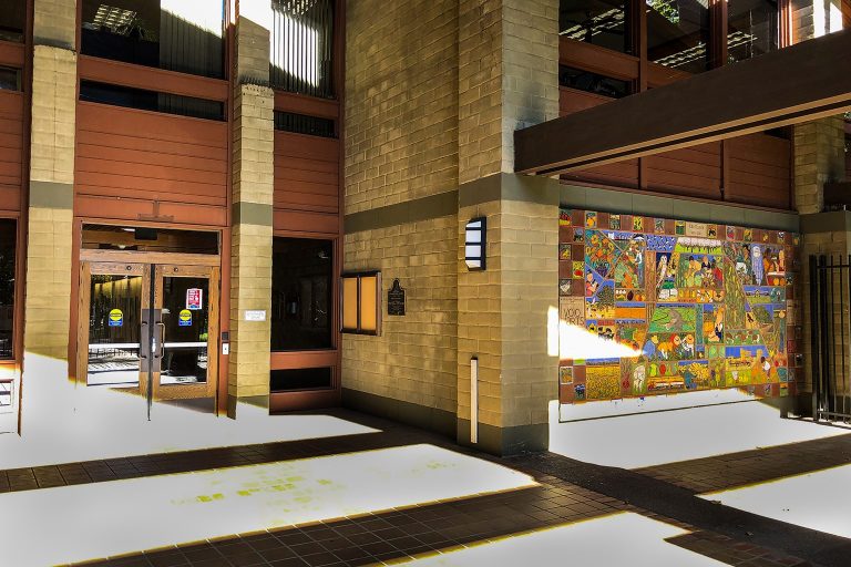 Sunlit brick building entrance with wooden doors on the left and a colorful mural displayed on the right wall.