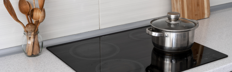 Stainless steel pot with lid on a glass electric cooktop, next to a jar of wooden utensils and cutting boards on a kitchen counter.
