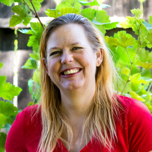 A middle-aged woman, VCE Board Member, with long blonde hair and a red shirt smiles before green leaves and a wooden fence.