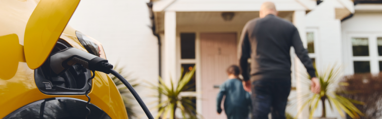 A yellow electric car is charging in a driveway while an adult and child walk toward the front door of a house.