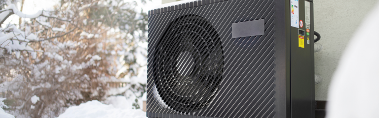 Outdoor heat pump unit in a snowy setting, mounted next to a building, with trees visible in the background.