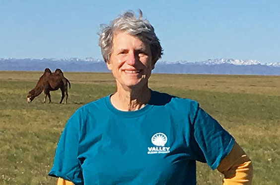 Smiling person in a blue Valley Clean Energy shirt stands in a grassy field with a camel grazing in the background.